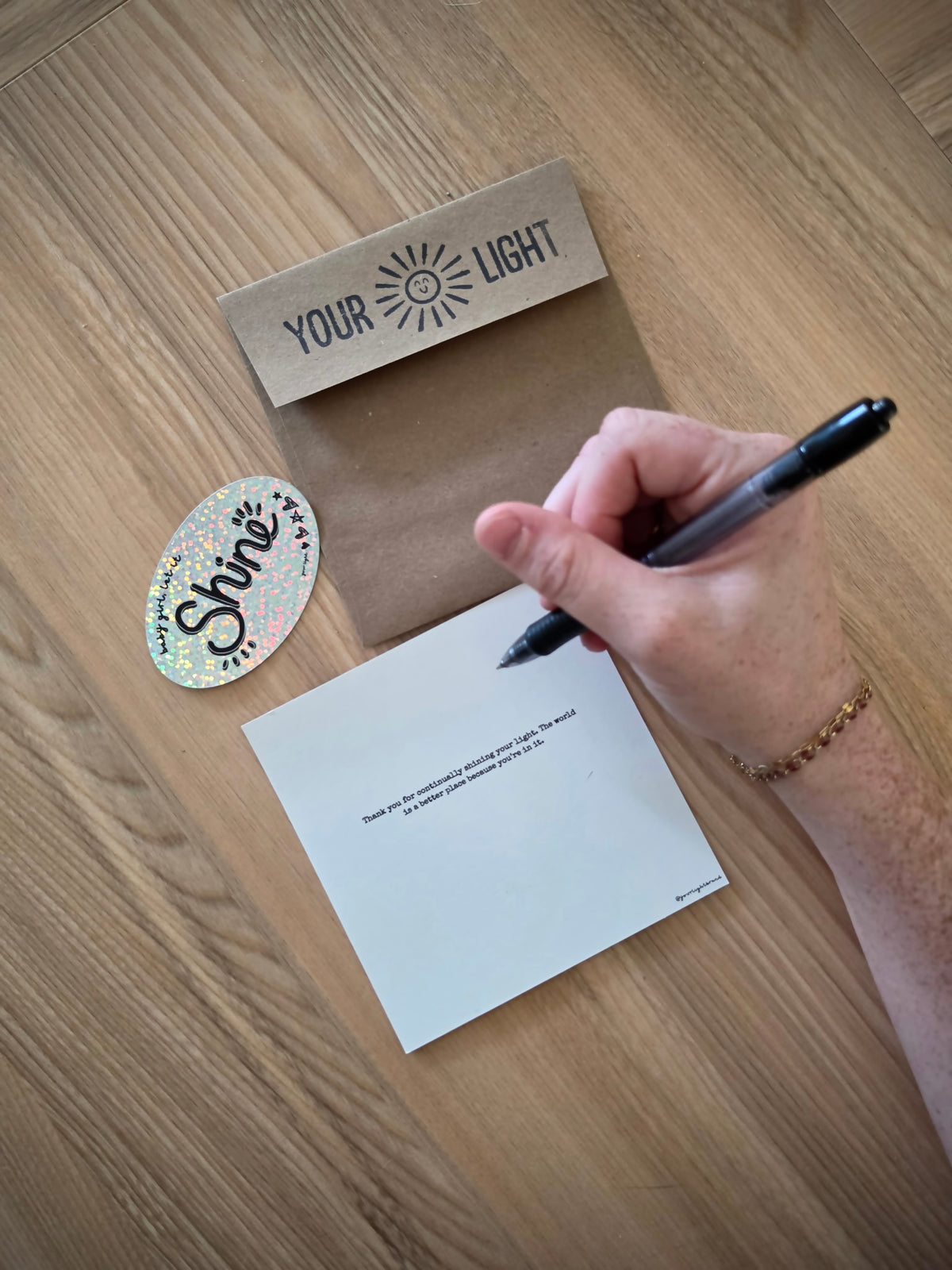 A hand holding a black pen writes on a white greeting card placed on a wooden table. The greeting card has the text 'Thank you for continually shining your light. The world is a better place because you're in it.' Nearby are a kraft envelope with the words 'YOUR LIGHT' and a small oval sticker that reads 'Shine'.