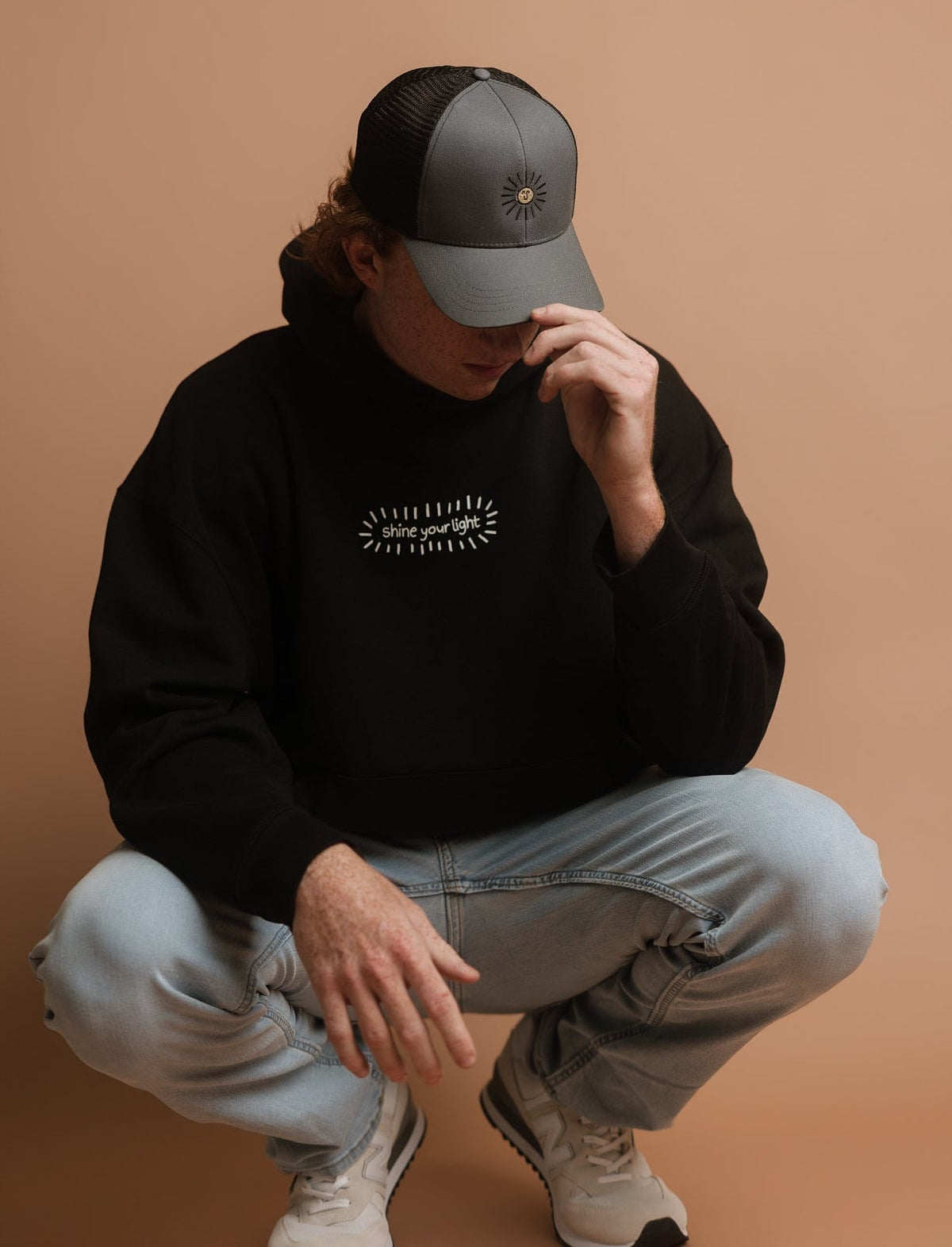 A person crouching against a solid warm tan studio background, wearing a dark gray trucker cap, a black sweatshirt with white text, and light blue jeans. The black sweatshirt features a graphic that says 'shine your light' in a handwritten font, surrounded by a burst of doodle lines.