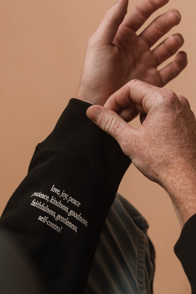 A close-up shot of a person's arm from above. The person is wearing a black long-sleeve sweatshirt with white printed text on the cuff, paired with light blue jeans against a solid warm beige background. The white printed text says 'love, joy, peace, patience, kindness, goodness, faithfulness, gentleness, self-control'.