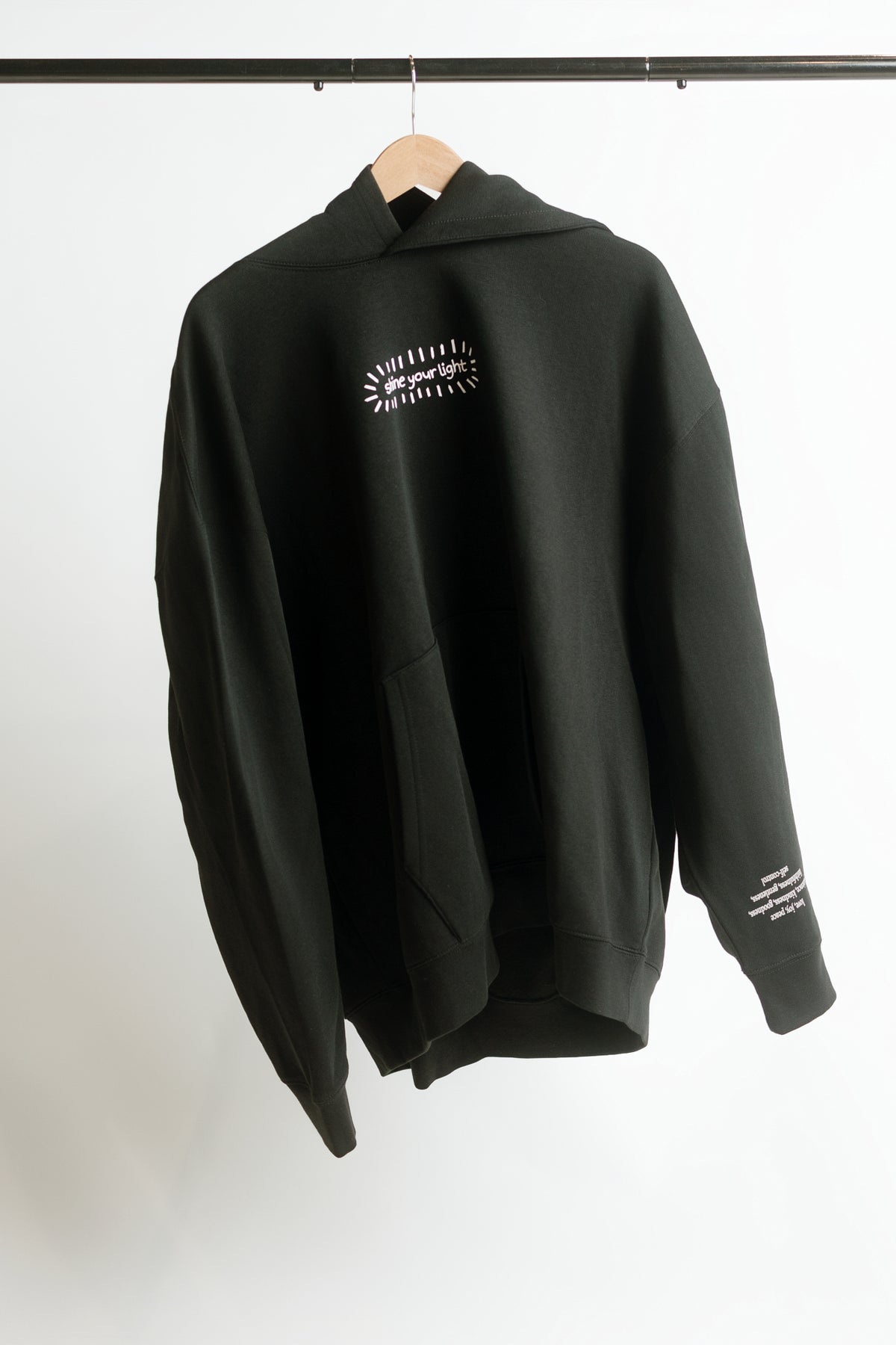 A black pullover hoodie hanging on a wooden hanger against a plain white wall. The hoodie features a small white graphic print on the left chest that says 'shine your light', and white text printed on the right sleeve.