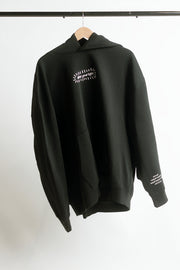 A black pullover hoodie hanging on a wooden hanger against a plain white wall. The hoodie features a small white graphic print on the left chest that says 'shine your light', and white text printed on the right sleeve.