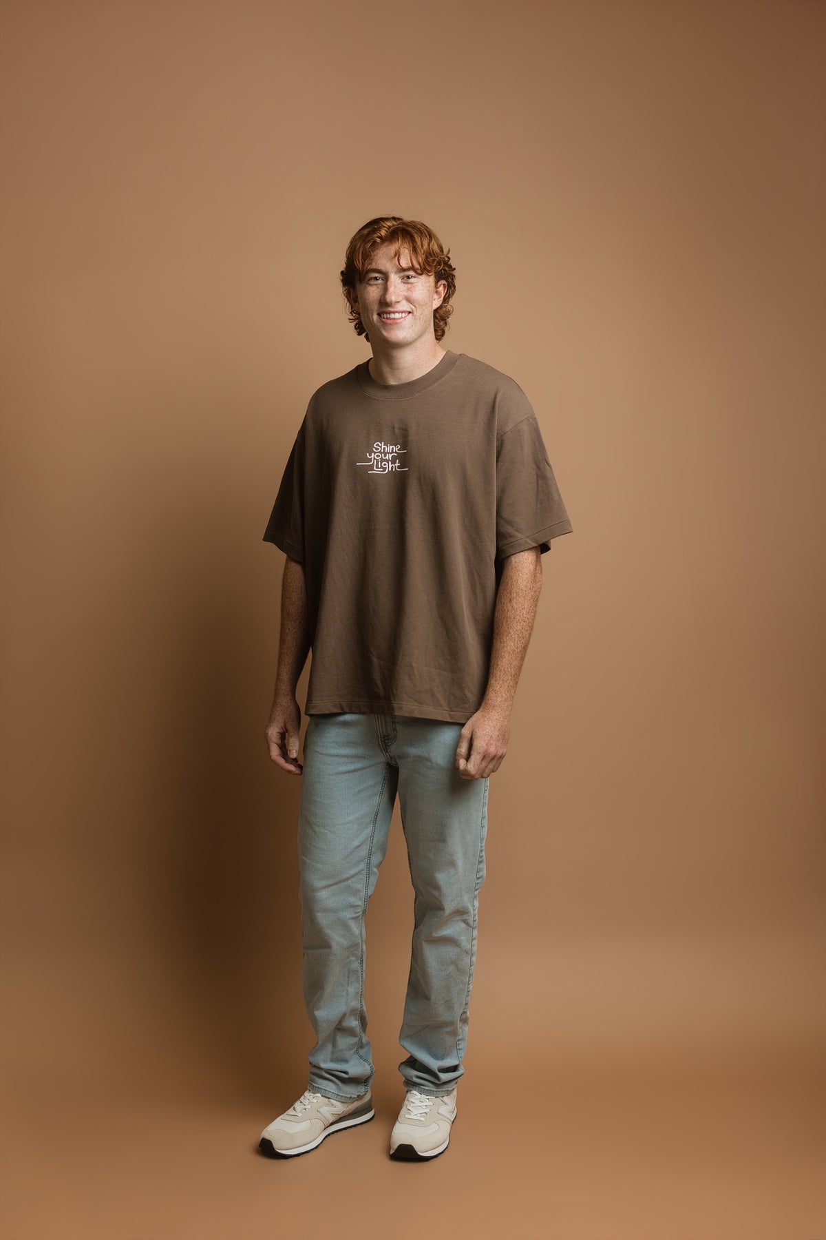 A young man with red hair stands against a solid brown studio backdrop, wearing an oversized brown t-shirt with a small white graphic on the chest, light blue jeans, and white sneakers. The graphic says 'Shine your light'.