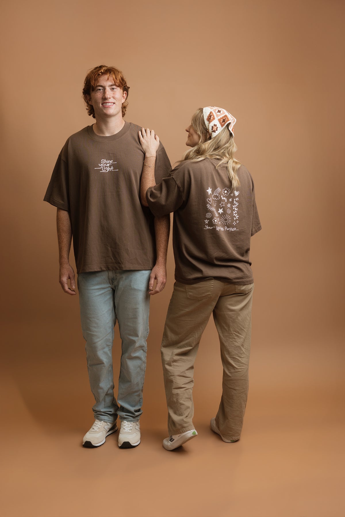 Two models wearing matching brown graphic t-shirts, standing against a solid brown studio backdrop. The model on the left faces forward, showing a small white graphic on the chest that says 'Shine your light', while the model on the right faces away, showing a larger white graphic with doodles of flowers, suns, and stars on the back.