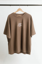 A brown, oversized, short-sleeve t-shirt with a white graphic print reading 'Shine your light' hanging on a wooden hanger against a white background.