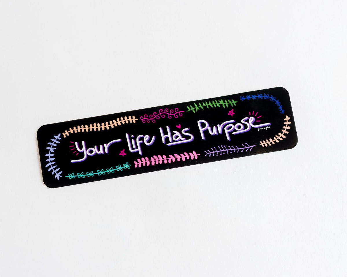 A rectangular black sticker with rounded corners featuring the text 'Your Life Has Purpose' in white hand lettering, surrounded by colorful doodle vines and flowers in pink, green, blue, and orange. The sticker is pictured on a white background.