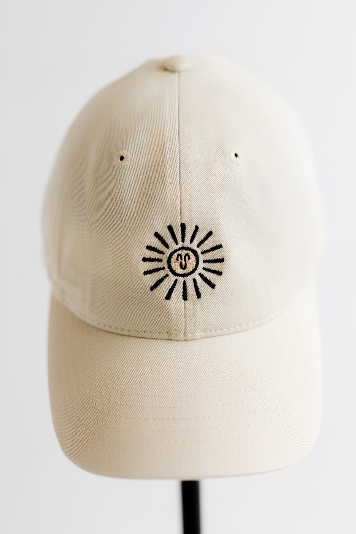 A cream-colored cotton baseball cap with a black and yellow embroidered doodle sun logo with a smiley face in the center, displayed on a black stand against a white background.