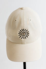 A cream-colored cotton baseball cap with a black and yellow embroidered doodle sun logo with a smiley face in the center, displayed on a black stand against a white background.