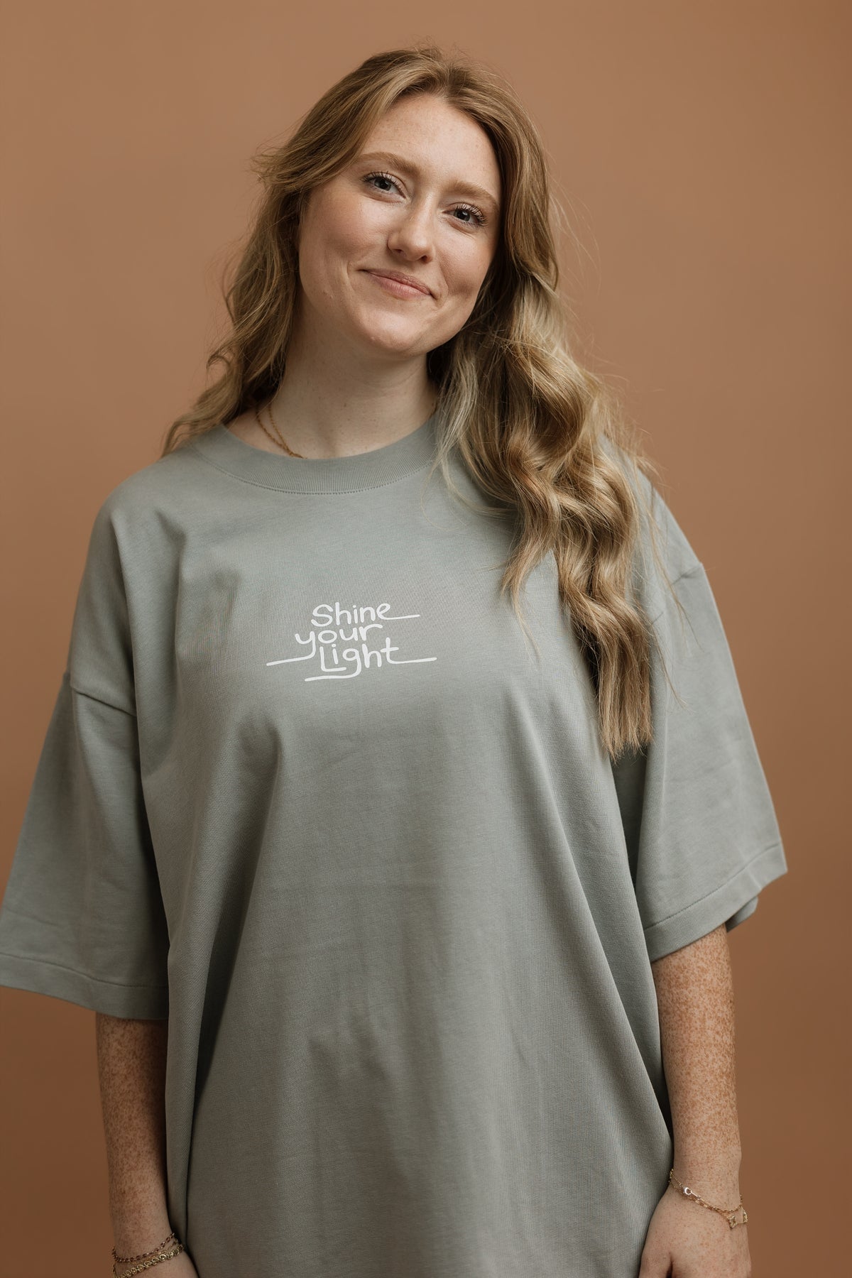A woman with long wavy blonde hair wearing an oversized gray t-shirt with the text 'Shine your Light' printed in white on the chest, posed against a solid warm brown studio background.