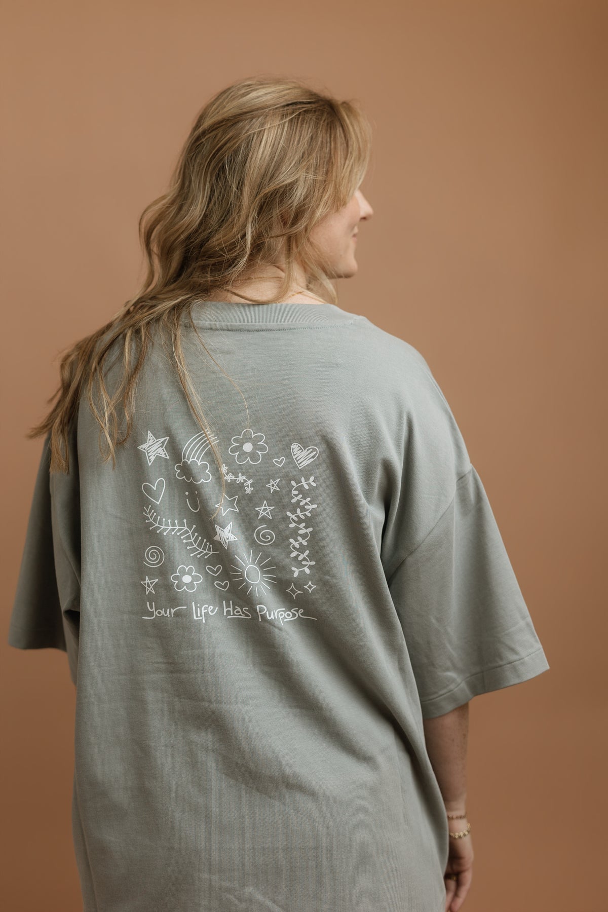 A woman with wavy blonde hair is shown from behind wearing an oversized gray t-shirt with a white hand-drawn style graphic on the back. The graphic features stars, hearts, flowers, and the text 'Your Life Has Purpose'.
