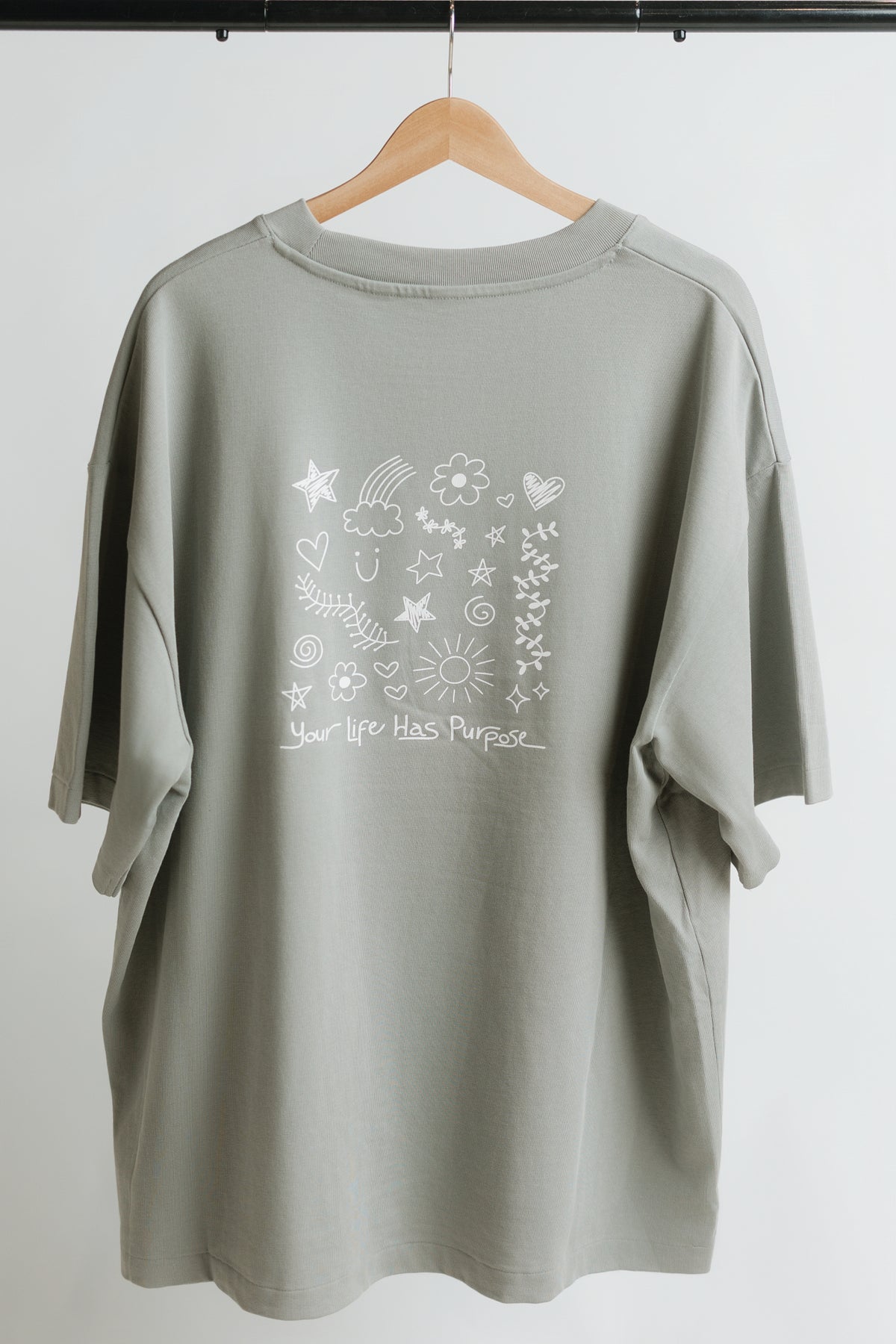 A gray, oversized short-sleeve t-shirt hanging on a wooden hanger against a white background. The back of the shirt features a white hand-drawn style graphic with stars, flowers, hearts, and the text 'Your life has Purpose' written in a handwritten font.