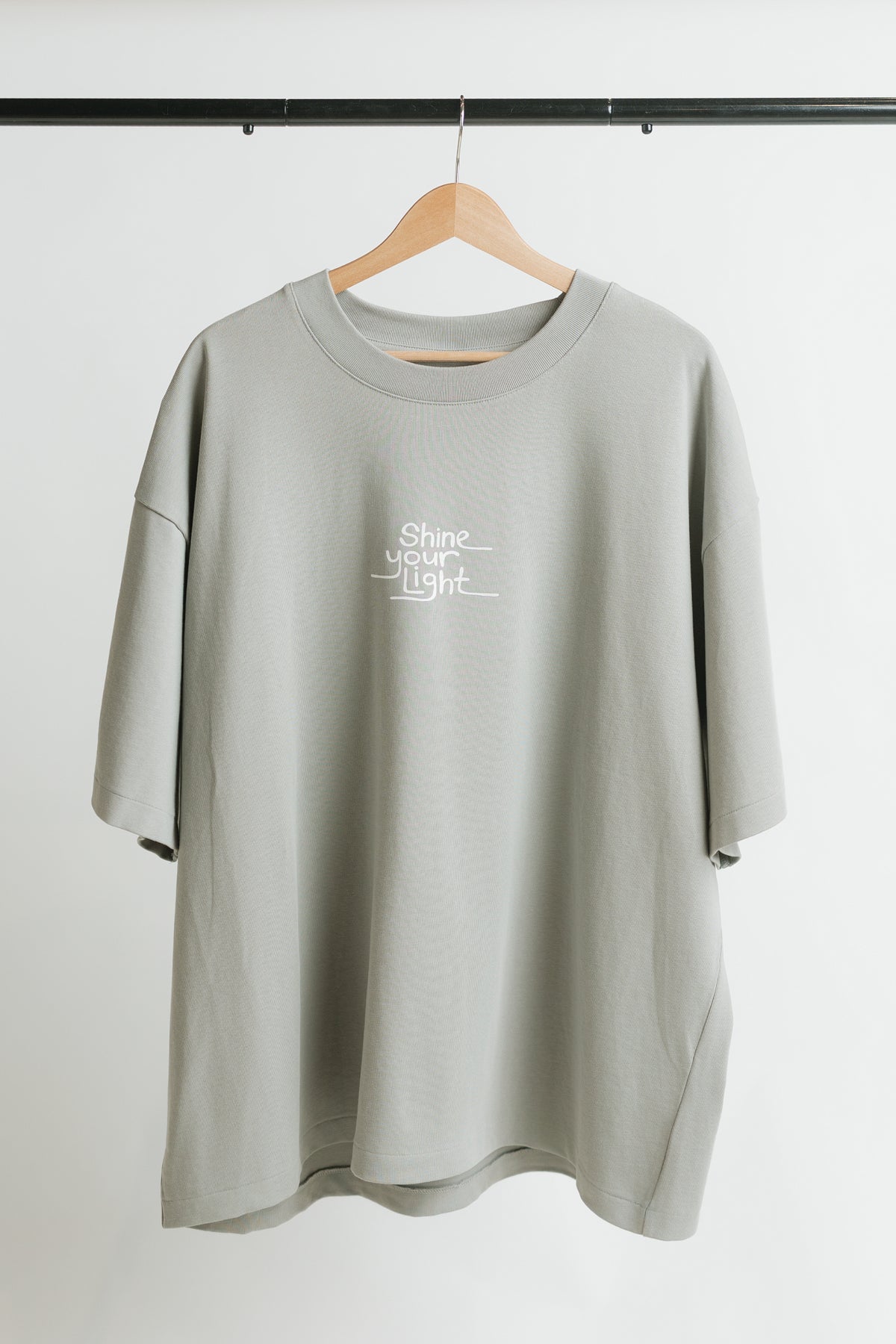 A light gray oversized short-sleeve t-shirt with a white graphic print reading 'shine your light' on the chest, hanging on a wooden hanger against a white wall.