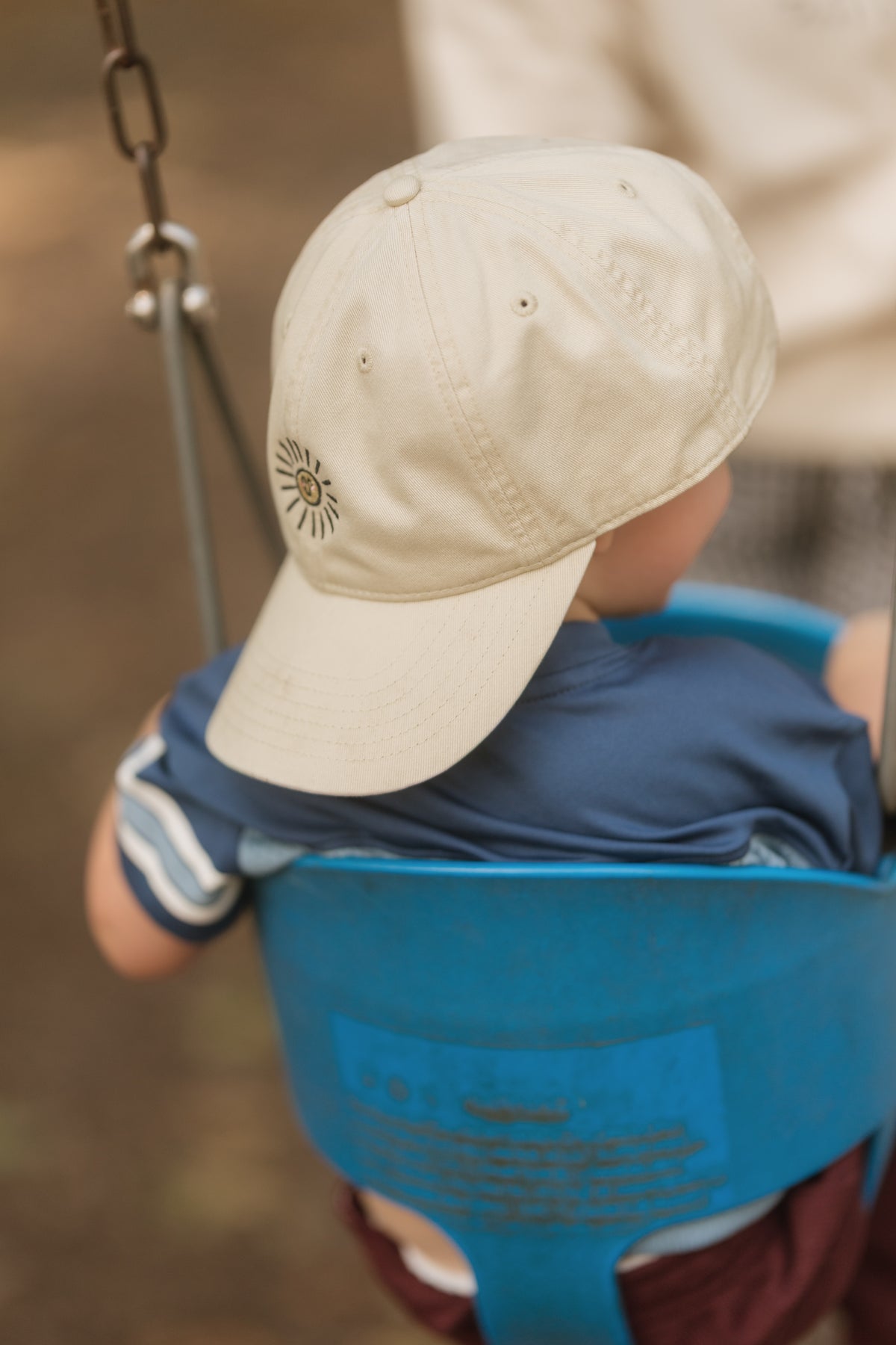 A child sitting in a blue swing seat viewed from behind, wearing a light beige baseball cap with a small embroidered sun doodle and a navy shirt.
