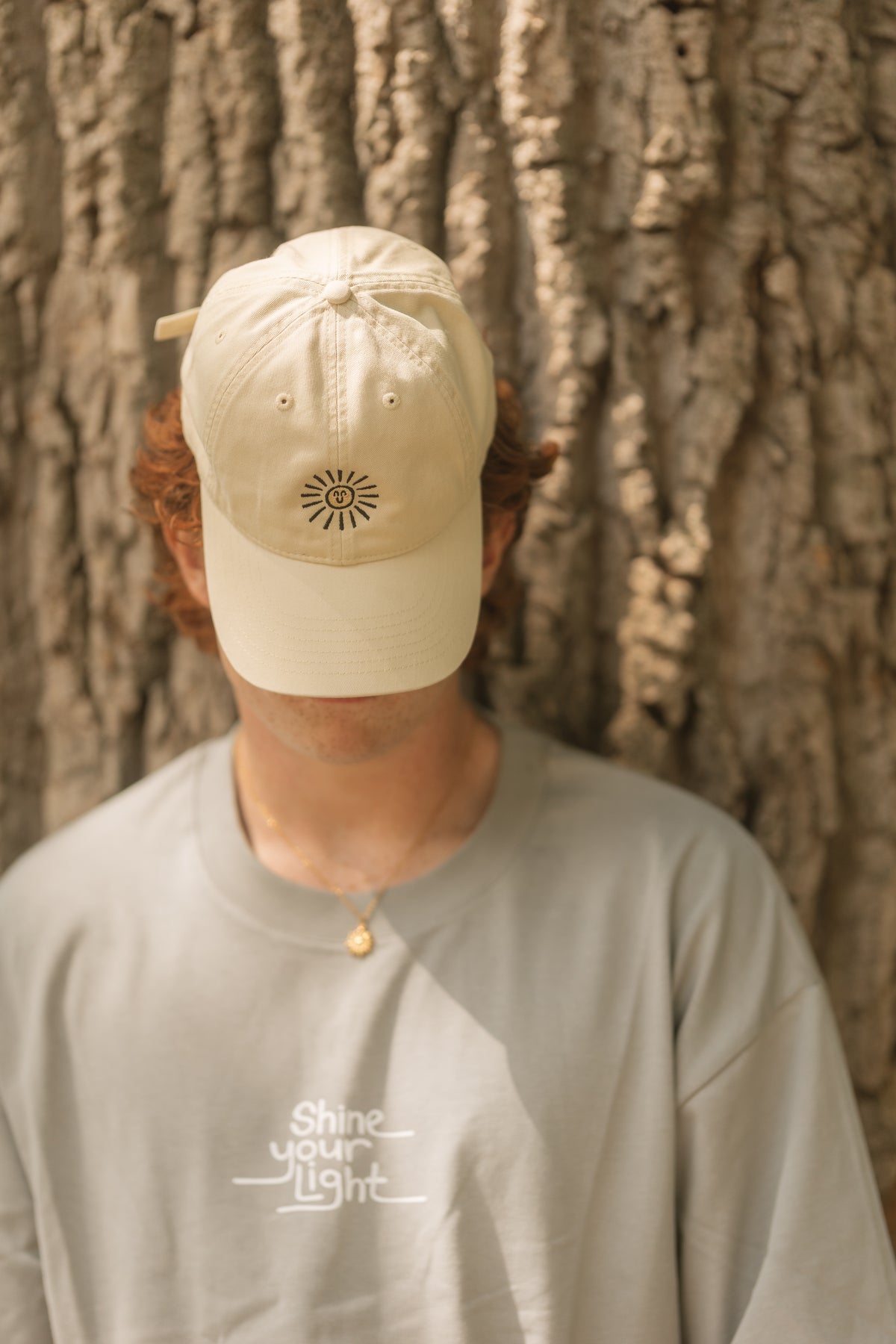 A person wearing a cream-colored baseball cap with an embroidered sun on the front, standing in front of a tree trunk. The person is also wearing a light gray t-shirt with 'shine your light' printed on the chest.