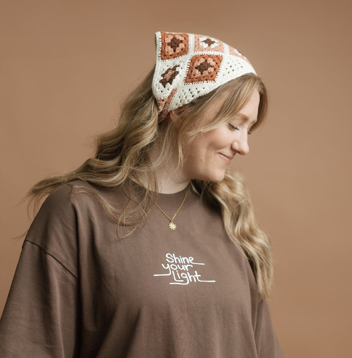 A woman wearing a brown oversized t-shirt with a white graphic print on the chest and a white crochet headband with brown patterns, posed against a solid brown studio background.