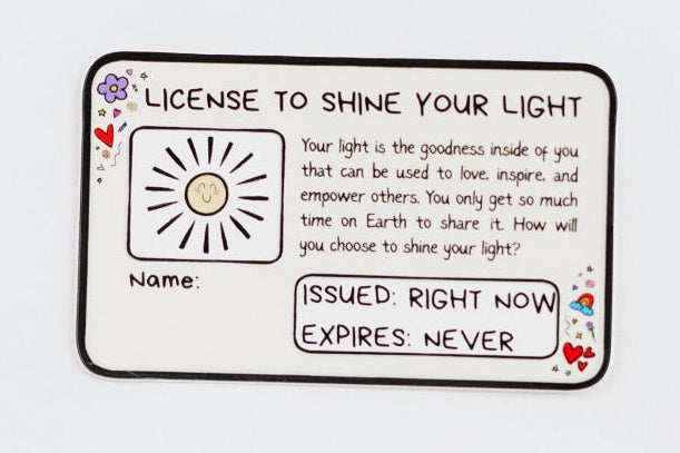 A styled flat-lay of various paper goods and stationery items on a clean white background, including a tag, a small card with a sun illustration and text, a sun sticker, a brown envelope and white greeting card, and a glittery oval sticker that says 'Shine'.
