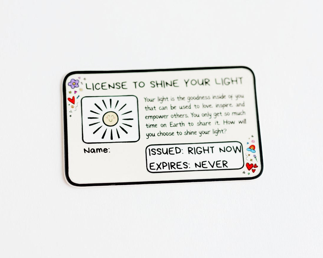 A rectangular white sticker with rounded corners and a black border and handwritten text, featuring a small sun illustration and decorative floral corner details, photographed on a plain white background. The sticker says 'License to shine your light' in a black handwritten font. The sticker is in the style of a drivers license, and features some text, including 'issued: right now' and 'expires: never'.