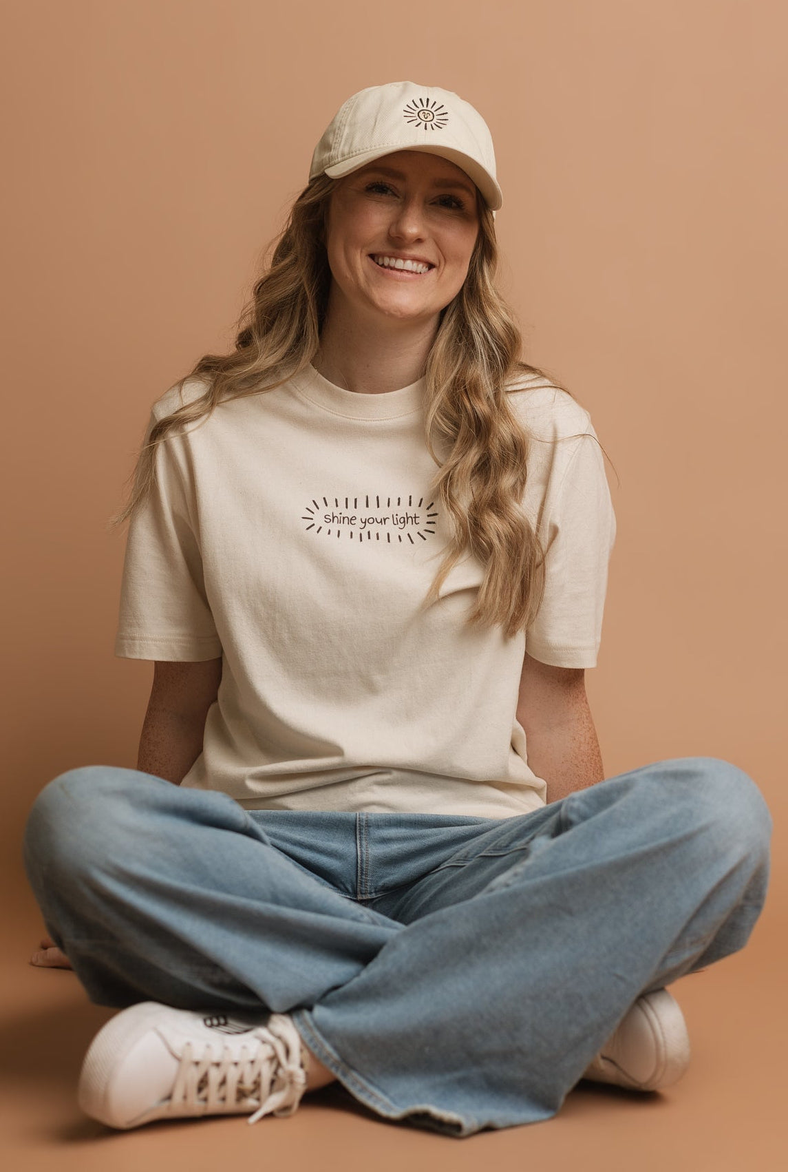 A woman sitting cross-legged on a solid tan studio background, wearing a cream-colored graphic t-shirt, a matching cream baseball cap, light blue jeans, and white sneakers. The graphic on the t-shirt says 'shine your light' in a black, handwritten font, and is surrounded by a burst of doodle lines.