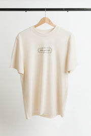 A cream-colored short-sleeve t-shirt hanging on a wooden hanger against a white wall. The shirt features a small black graphic print on the center chest with the words 'shine your light' in a handwritten font, surrounded by a burst of doodle lines