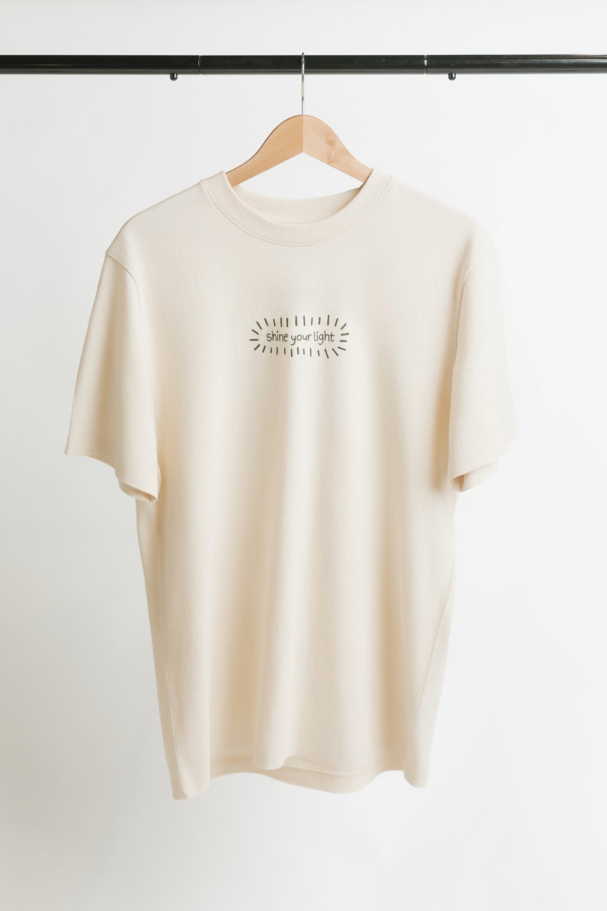 A cream-colored short-sleeve t-shirt hanging on a wooden hanger against a white wall. The shirt features a small black graphic print on the center chest with the words 'shine your light' in a handwritten font, surrounded by a burst of doodle lines