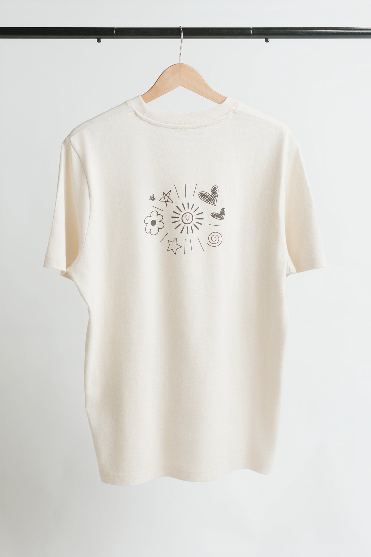 A cream-colored short-sleeve t-shirt hanging on a wooden hanger against a white wall, featuring a black line art graphic of a sun, butterflies, and flowers in the center of the back of the shirt.