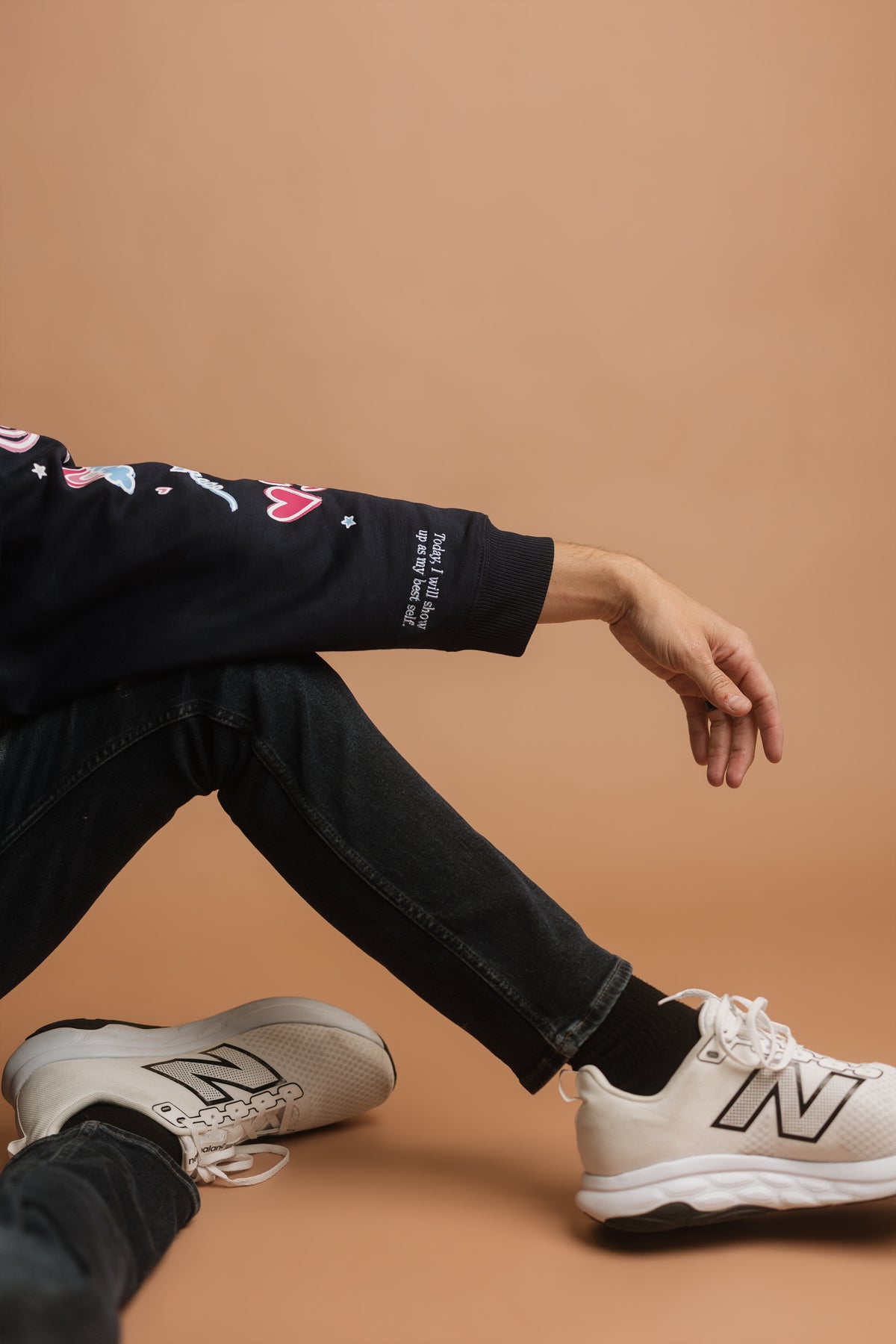 A person sitting with one knee up, wearing a navy long-sleeve graphic sweatshirt, black jeans, and white sneakers against a solid tan background. The hem of the sleeve of the sweatshirt says 'today I will show up as my best self'.