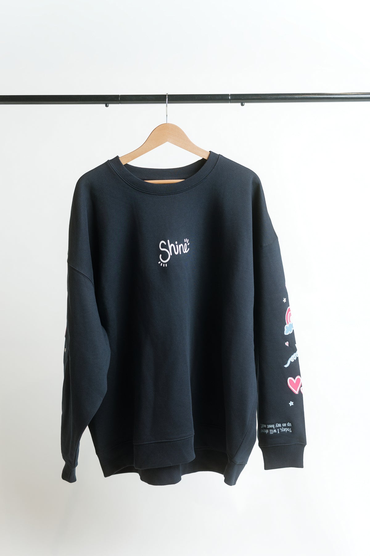 A navy crewneck sweatshirt hanging on a wooden hanger against a white wall, featuring a white 'Shine' graphic on the chest, colorful doodle sleeve prints, and white text on the hem of the sleeve.