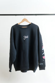 A navy crewneck sweatshirt hanging on a wooden hanger against a white wall, featuring a white 'Shine' graphic on the chest, colorful doodle sleeve prints, and white text on the hem of the sleeve.