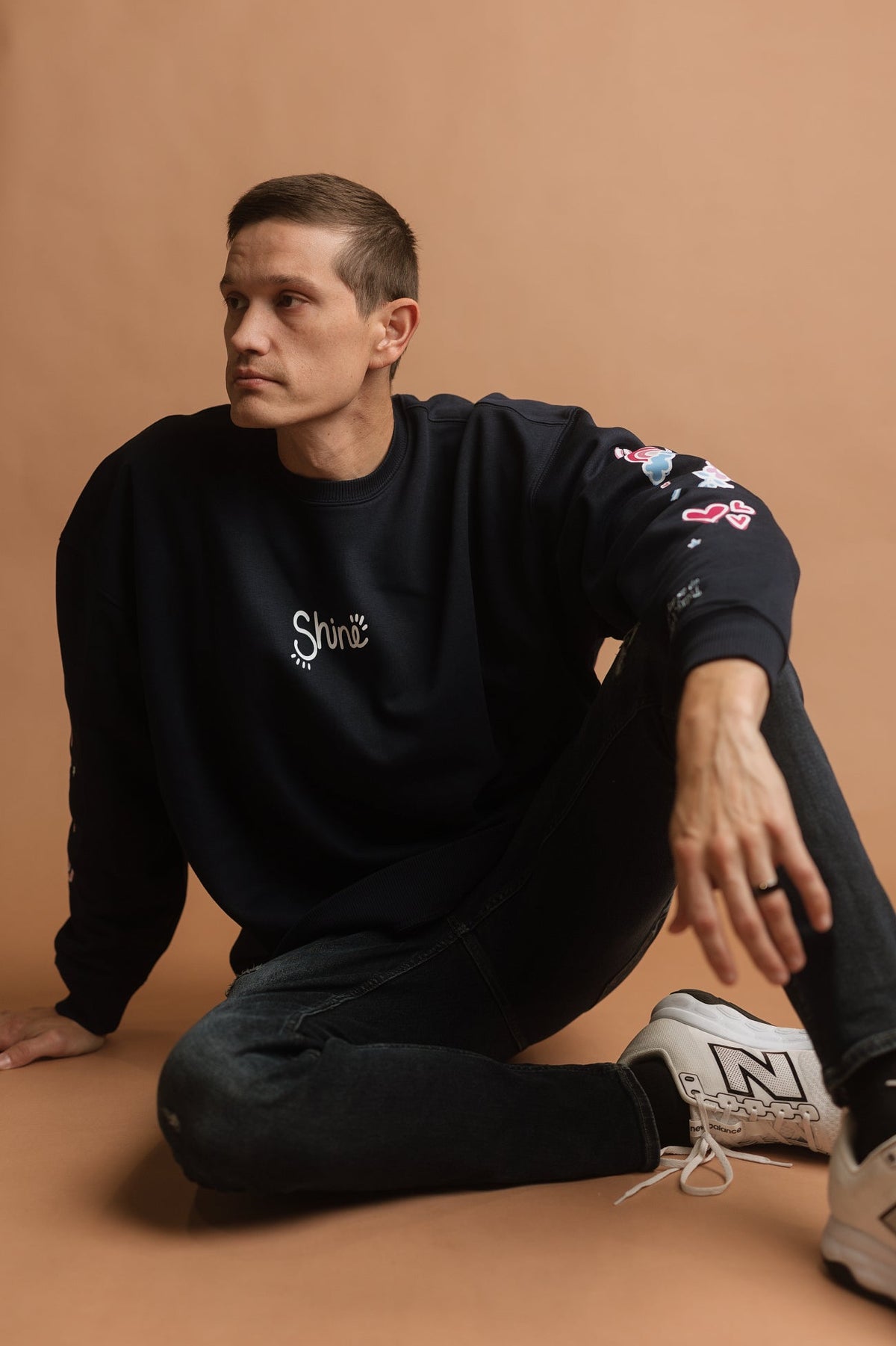 A man wearing a navy crewneck sweatshirt with a white 'Shine' logo on the chest and colorful doodles on the sleeve, paired with dark wash jeans and white sneakers, sitting against a solid tan studio background.