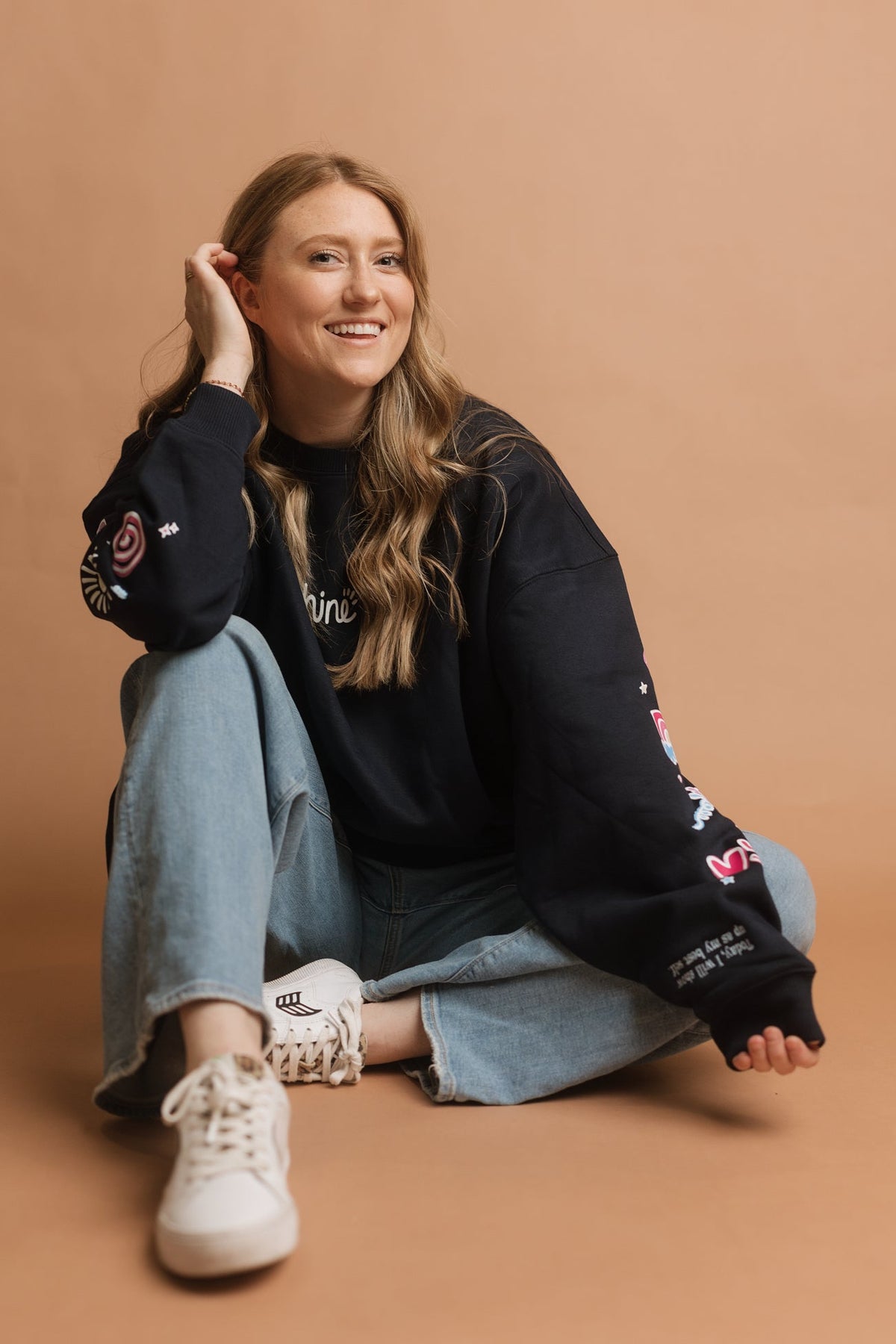 A woman sitting on a warm brown studio floor wearing a navy crewneck sweatshirt with colorful doodle details on the sleeves, light blue jeans, and white sneakers.
