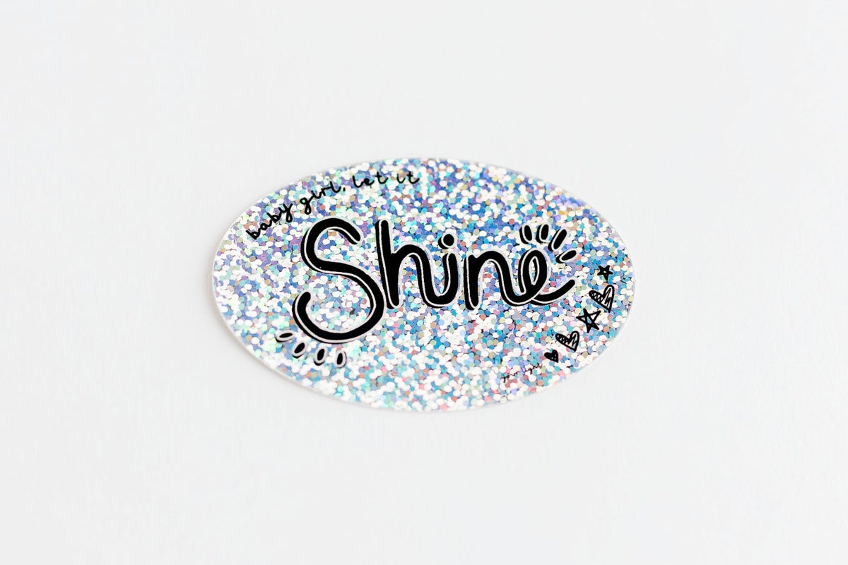 An oval sticker with a holographic glitter background and the word 'baby girl, let it Shine' in black script, shown on a plain white background.