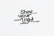 A die-cut sticker with the phrase 'Shine your Light' in black hand lettering on a white background, shown against a clean white backdrop.