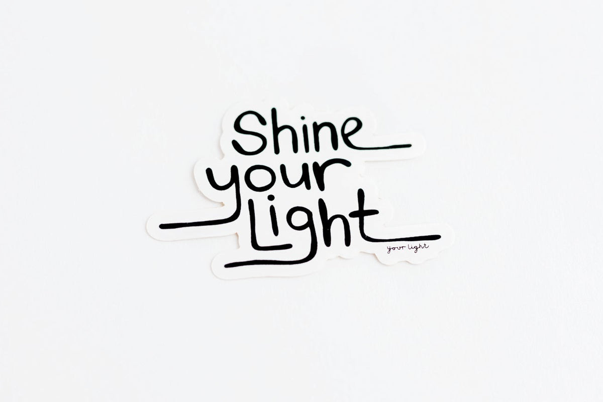 A die-cut sticker with the phrase 'Shine your Light' in black hand lettering on a white background, shown against a clean white backdrop.