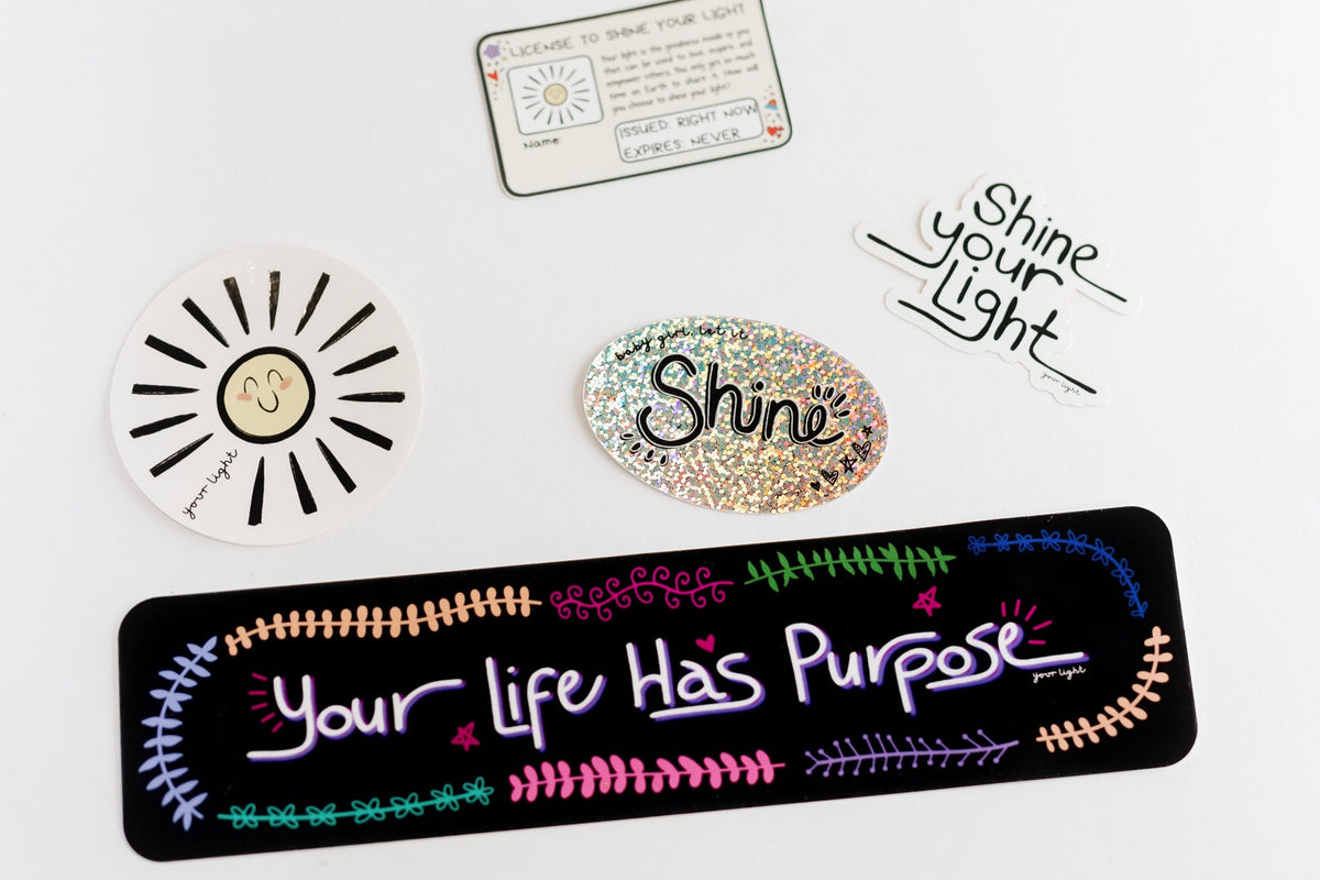 A flat-lay arrangement of several inspirational stickers and a small card on a white background. The items include a rectangular black sticker with colorful handwritten-style text reading 'Your Life Has Purpose,' a white sun face sticker, a glittery oval sticker reading 'Shine,' a handwritten-style sticker reading 'Shine Your Light,' and a small card with text and a sun doodle.