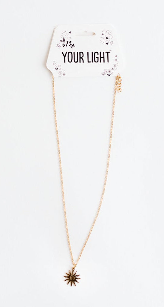A delicate rose gold necklace with a small swirly starburst pendant, displayed on a white card that reads 'YOUR LIGHT' against a solid white background.