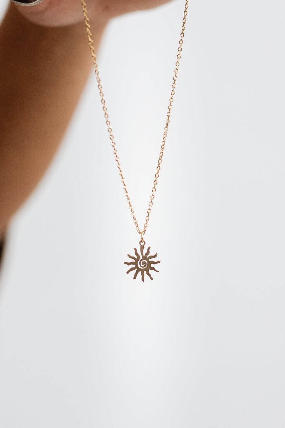 A gold chain necklace with a swirly sunburst pendant is held up by a hand with pink nail polish against a plain white background.