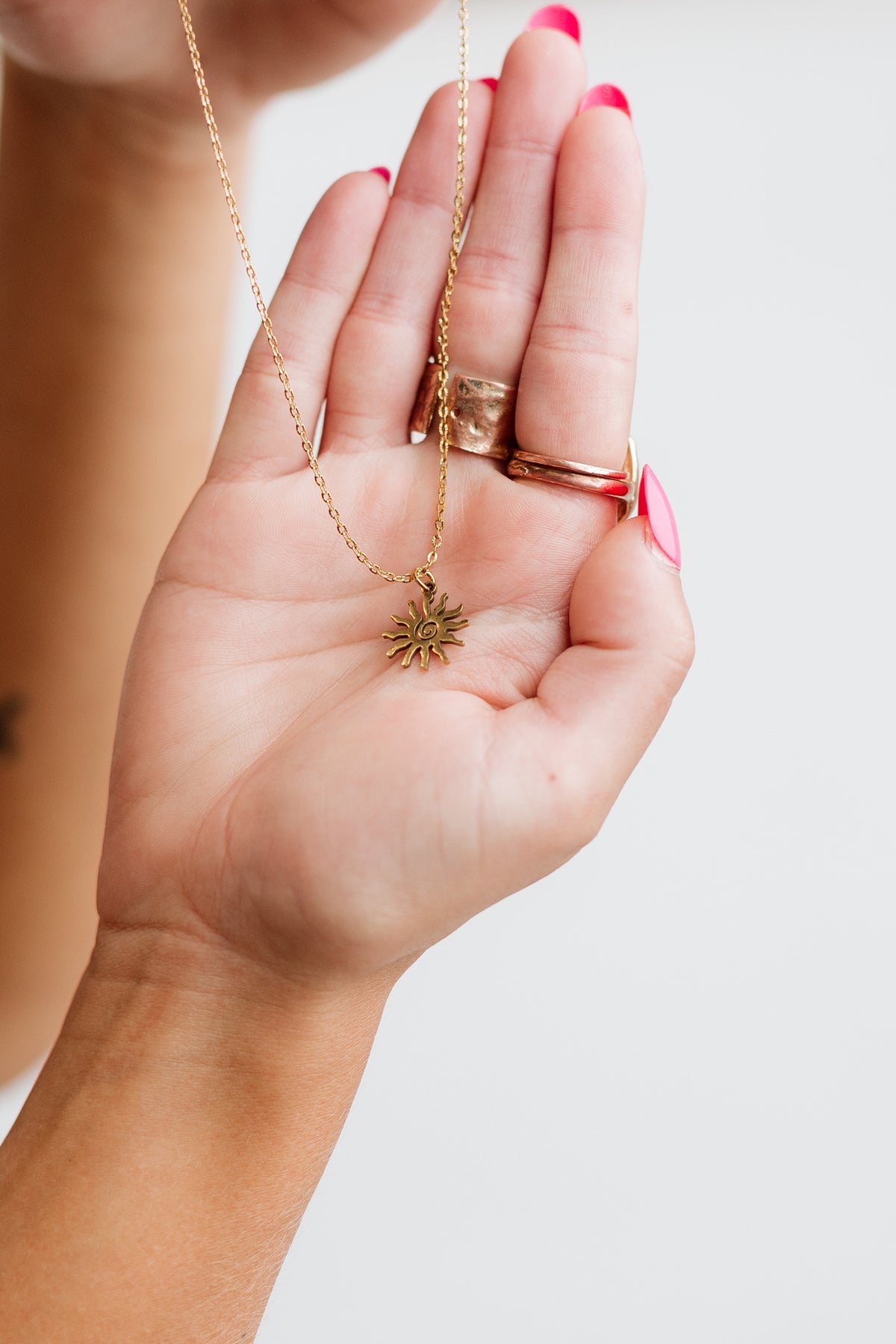 A close-up of a person's hand holding a delicate gold chain necklace with a small swirly sunburst pendant, against a plain white background.