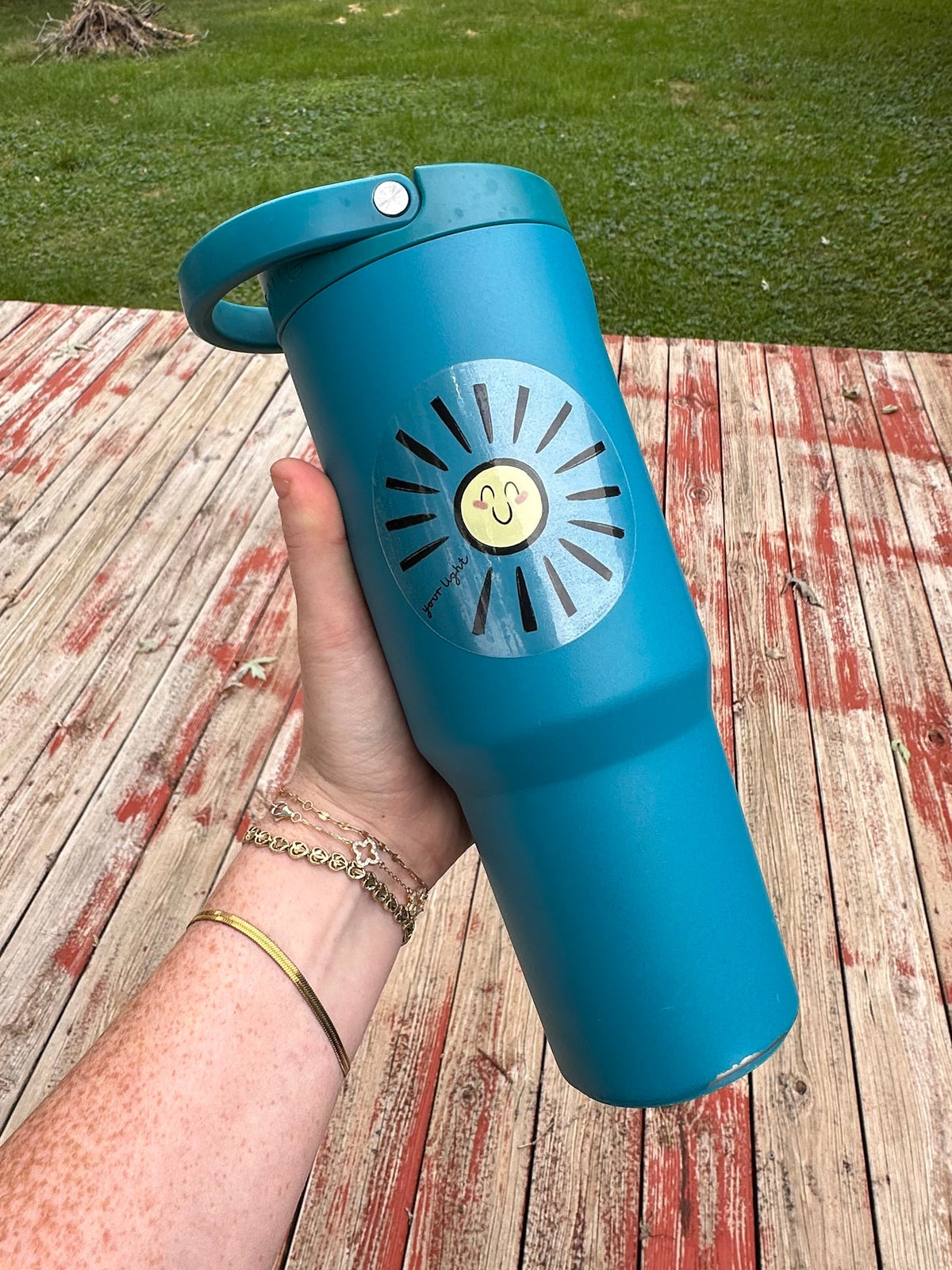A teal insulated tumbler with a doodle smiling sun sticker, held by a person's hand on a weathered wooden deck with a grassy backyard in the background.